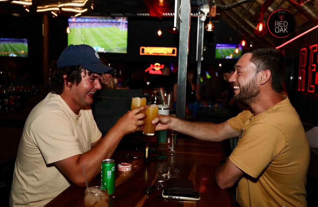 Two guests toasting beers with a rugby match on the screen behind them.