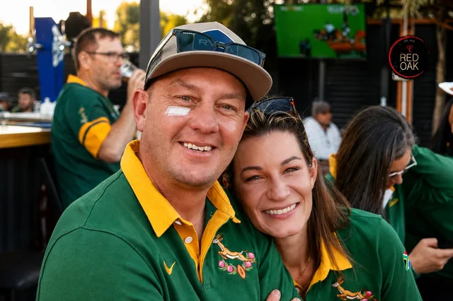A smiling couple in matching Springbok jerseys with a rugby match on the screen behind them.