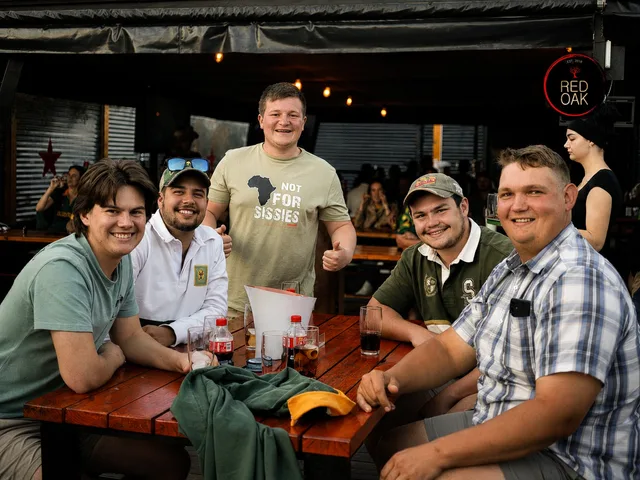 Five friends at a wooden picnic table with drinks, one in a Not For Sissies shirt.
