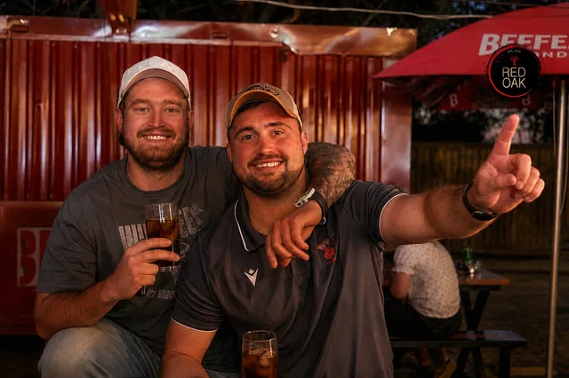 Two friends with drinks outside near a red shipping container, one pointing at the camera.