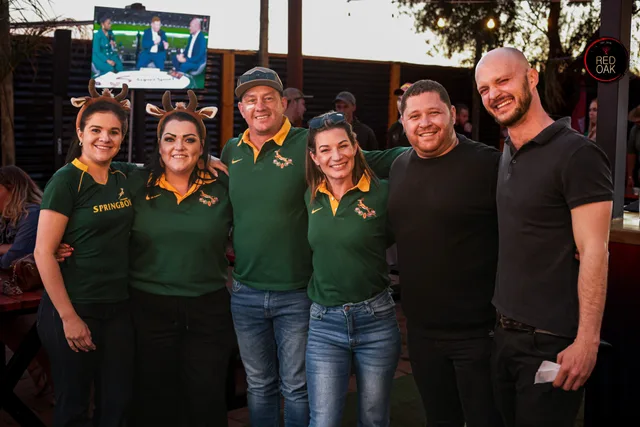 A group of friends in Springbok jerseys and antlers, standing together with a rugby match on the screen.
